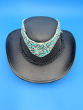 A black leather cowboy hat with a wide brim, featuring a black braided band and a turquoise and black patterned fabric band around the crown, displayed on a solid blue background.