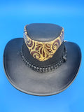 A black faux-leather wide-brim hat with a decorative patterned band in gold, brown, and black, set against a solid blue background.